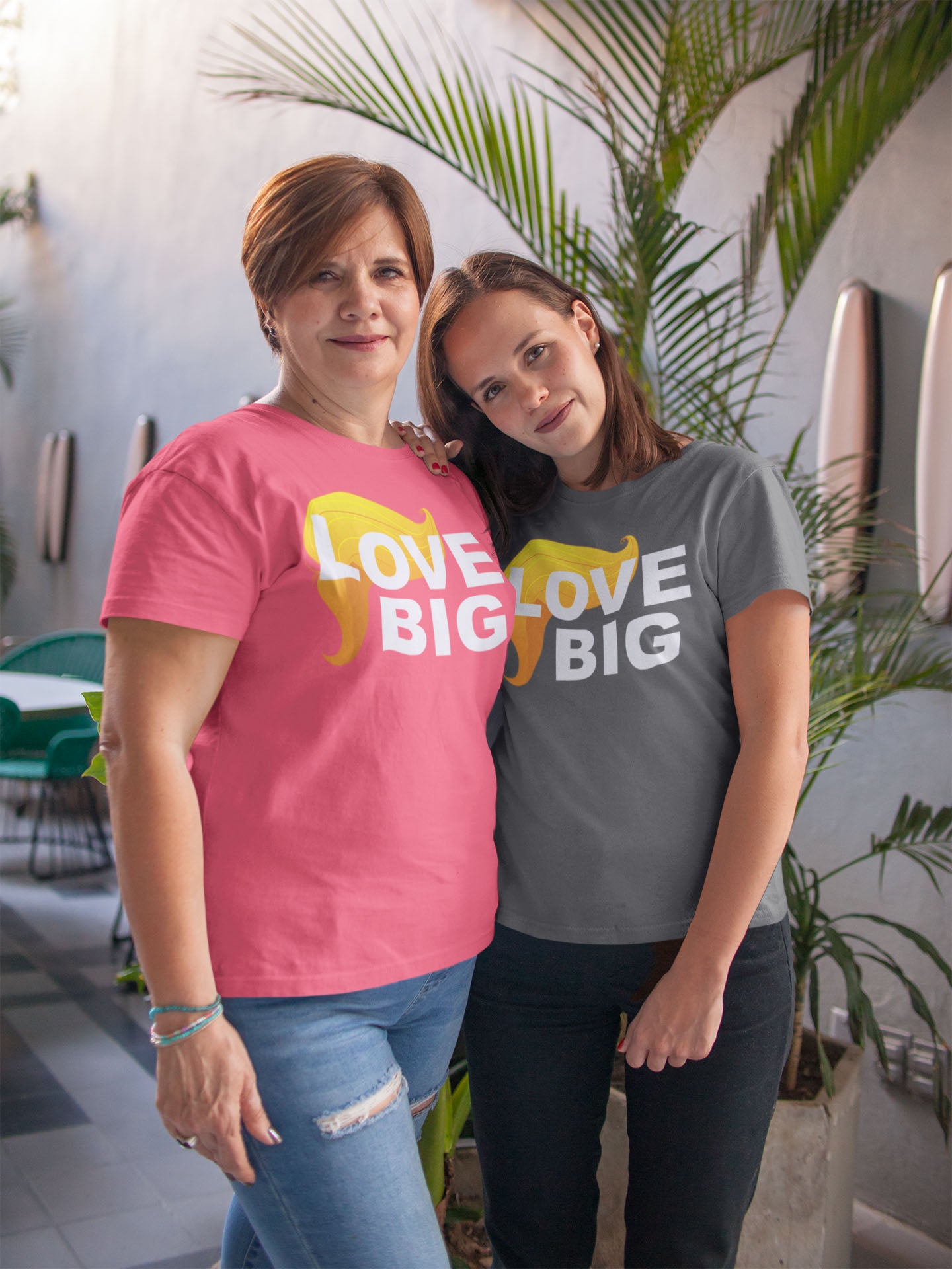 Two women wearing Love Big With Hair 2 Trump Humor Statement Tee shirts, showcasing vibrant political humor apparel.